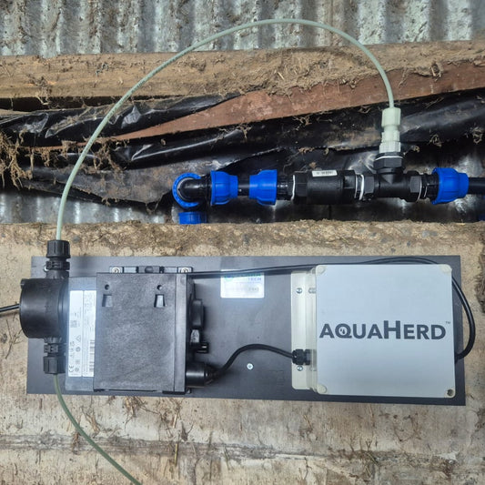 AQUAHerd automatic mineral dosing system mounted on a wooden wall, featuring a compact control unit connected to water and mineral lines to deliver liquid minerals through the herd’s drinking water.