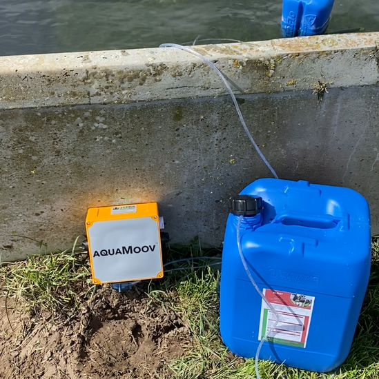AquaMoov battery-operated mobile mineral dosing system positioned beside an outdoor concrete water trough. The orange unit connects to a blue container of liquid minerals, automatically dosing minerals into the herd’s drinking water.