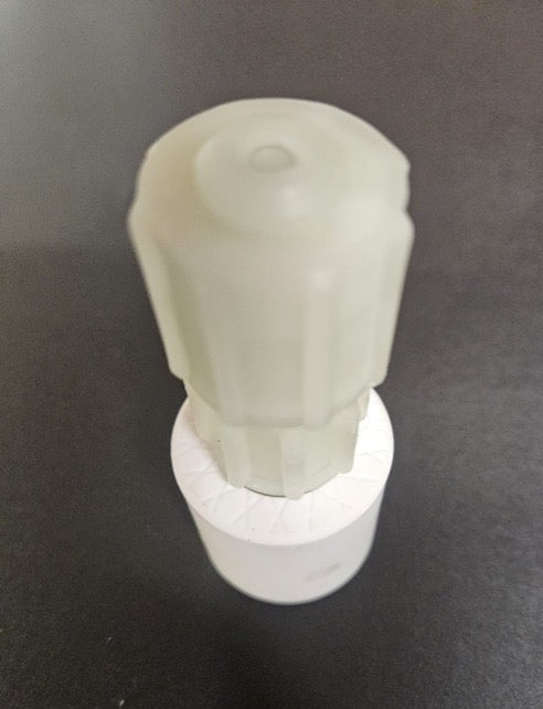 A translucent frosted white plastic cap or nozzle assembly mounted on a white cylindrical base, photographed on a dark gray surface.